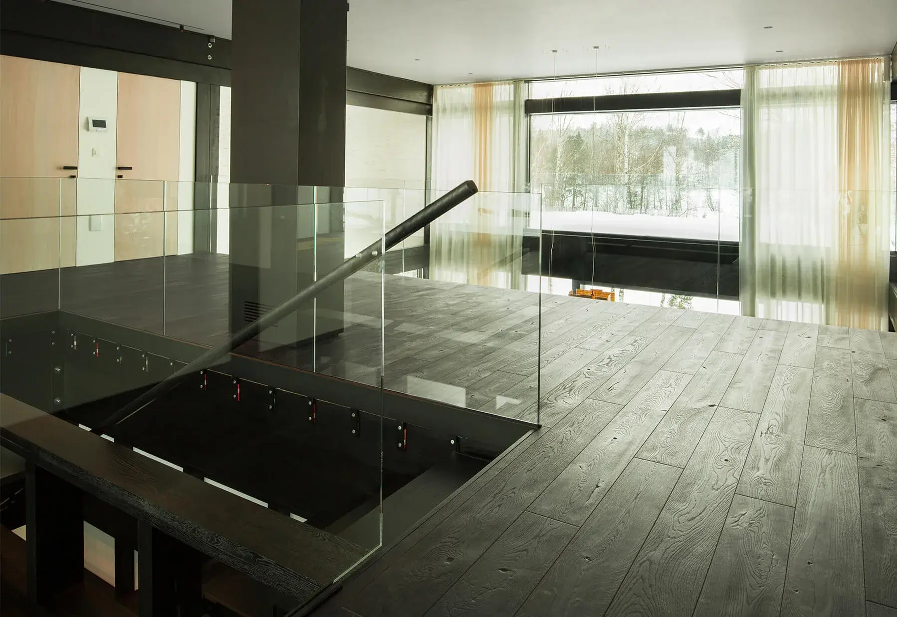 Moderní otevřený interiér s černou dubovou podlahou kolekce Ink, skleněným zábradlím a výhledem do zasněžené krajiny přes velkoformátová okna, doplněný textem o variabilitě odstínů černé.