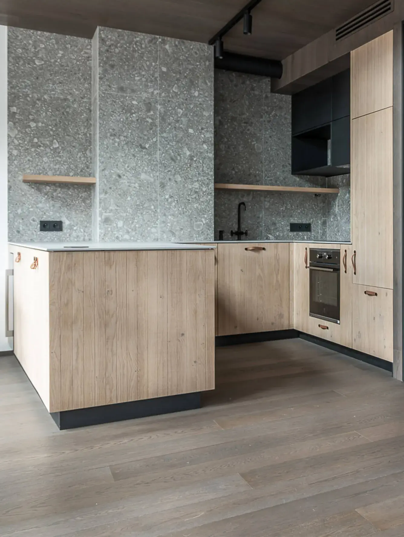 Moderní kuchyně v přírodním stylu s dřevěnou podlahou a ladícími čelními plochami skříněk s vertikálním frézováním, které doplňuje šedý kamenný obklad stěn s texturou terazza a minimalistické černé doplňky.