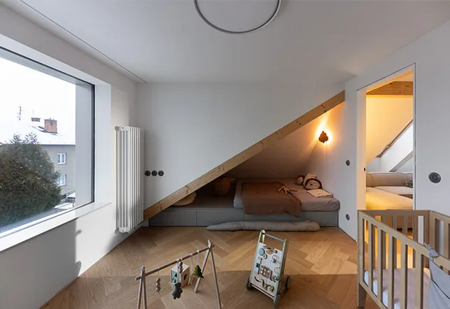 Moderní dětský pokoj ve střešním bytě v Olomouci s dubovou podlahou položenou ve vzoru herringbone, šikmou stěnou s vestavěným lůžkem a velkým oknem s výhledem na zasněžené střechy.