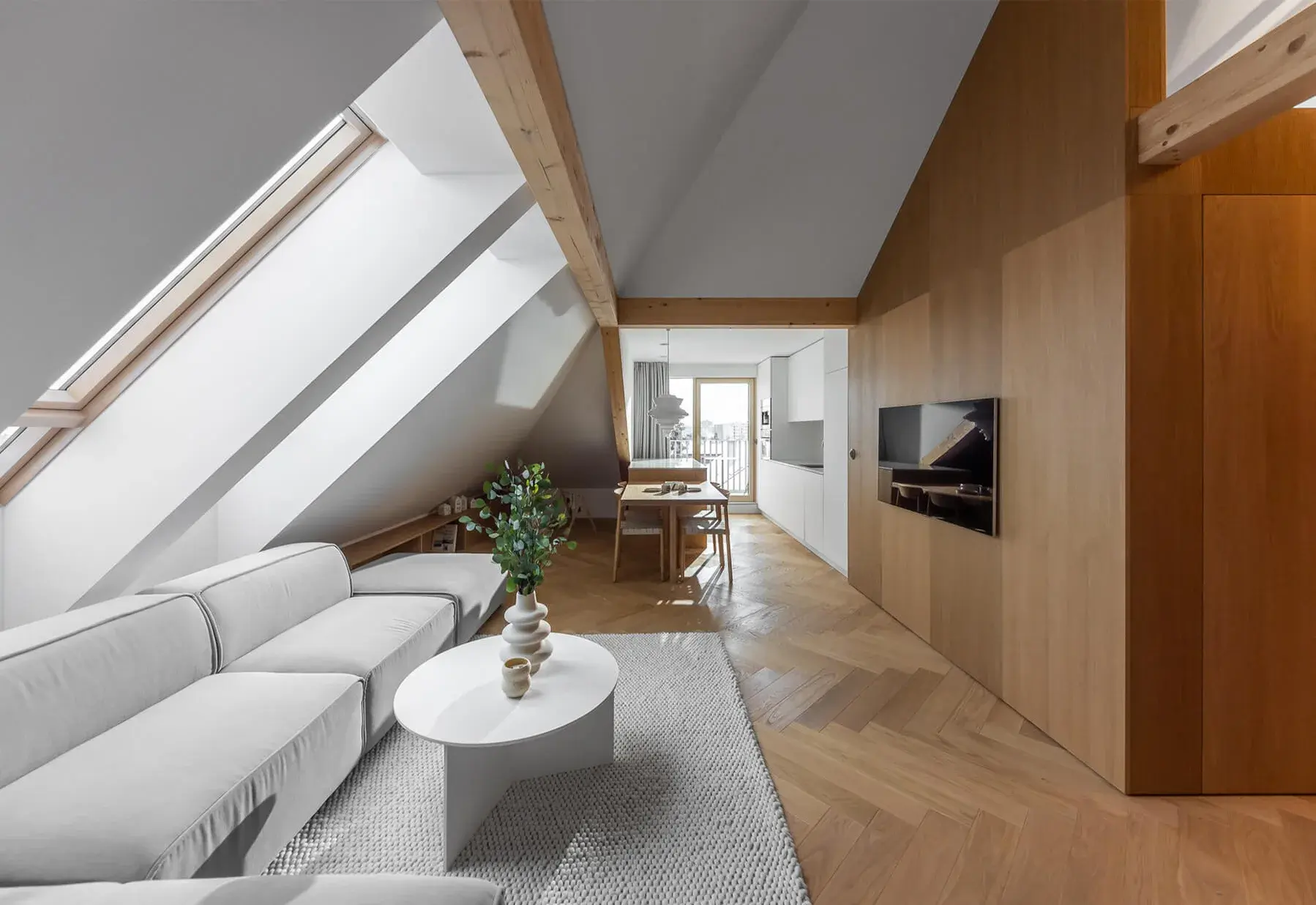 Moderní podkrovní interiér s dubovou podlahou položenou ve vzoru herringbone, doplněný masivním dřevěným obložením stěn a bílou sedací soupravou pod střešními okny.
