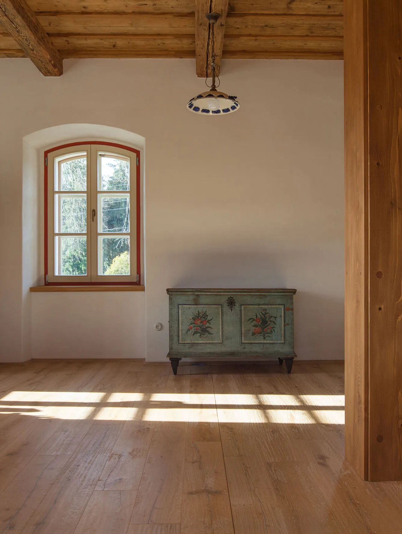 Rustikální interiér s dřevěnou podlahou v přírodním odstínu, kterému dominuje historická malovaná truhla u bílé stěny pod klenutým oknem, doplňující tradiční atmosféru s masivními stropními trámy.