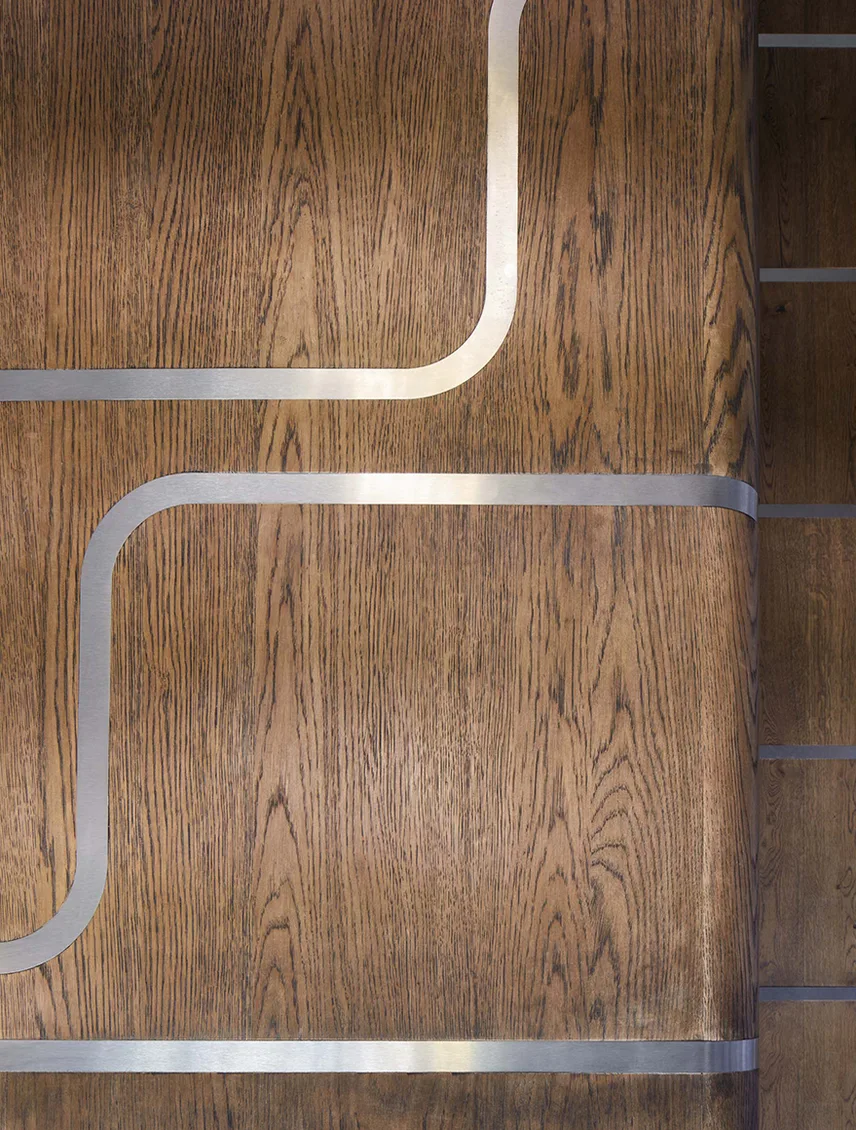 Detailní pohled na dřevěný schodišťový stupeň v sytém hnědém dekoru Hazel s dekorativním kovovým lemováním zapuštěným do povrchu dřeva.