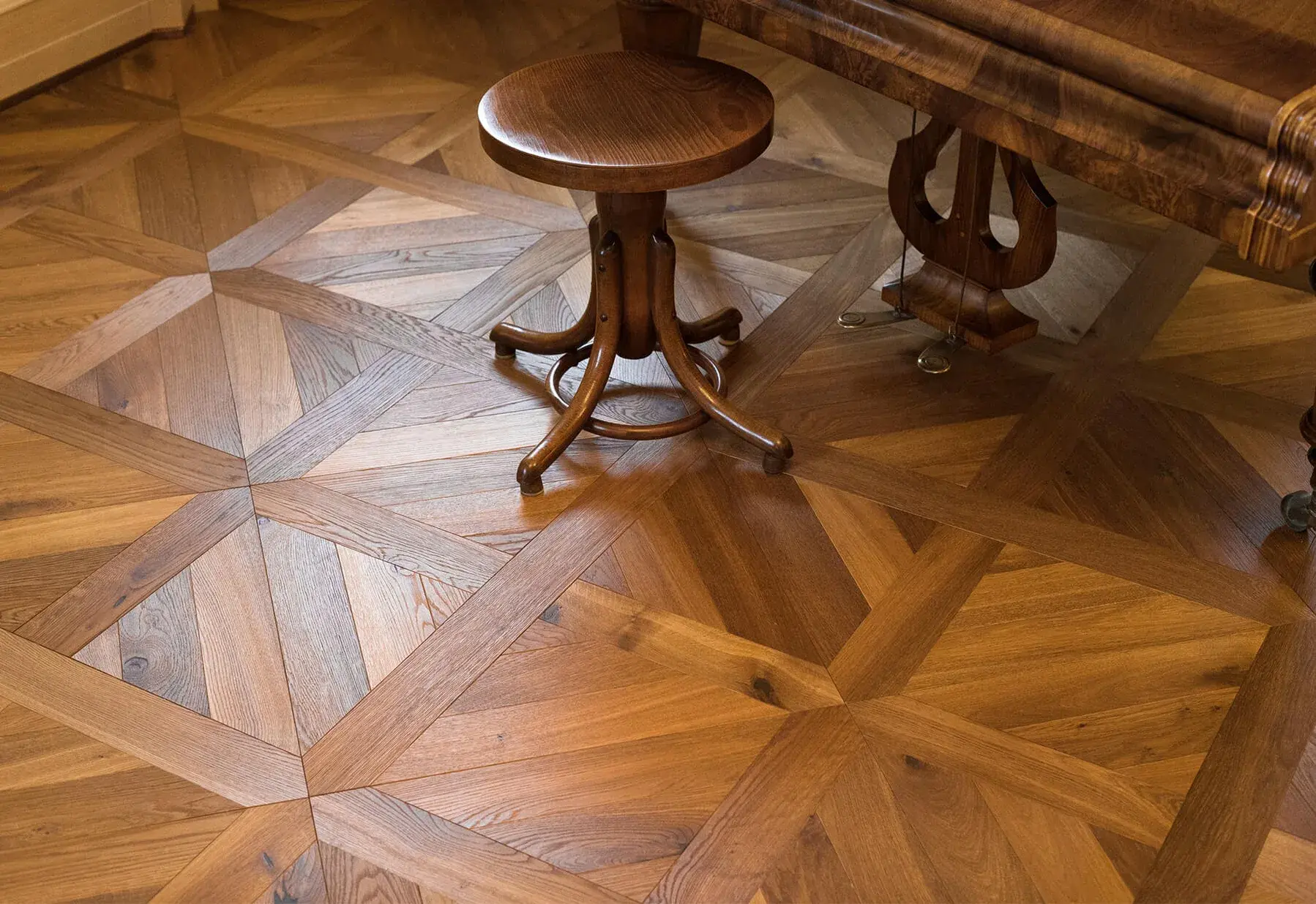 Detailní záběr na historickou dřevěnou podlahu s kazetovým vzorem v bohatém hnědém dekoru Hazel, doplněnou dobovým klavírem a dřevěnou stoličkou.