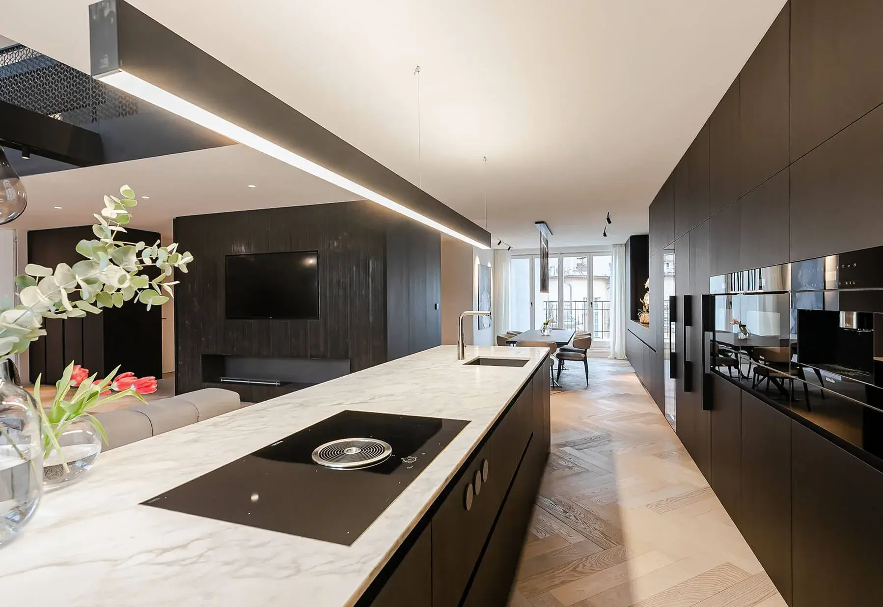 Moderní minimalistický interiér kuchyně a jídelny v luxusním mezonetovém bytě s bílou mramorovou pracovní deskou, tmavým nábytkem a dubovou podlahou položenou do vzoru Heringbone.