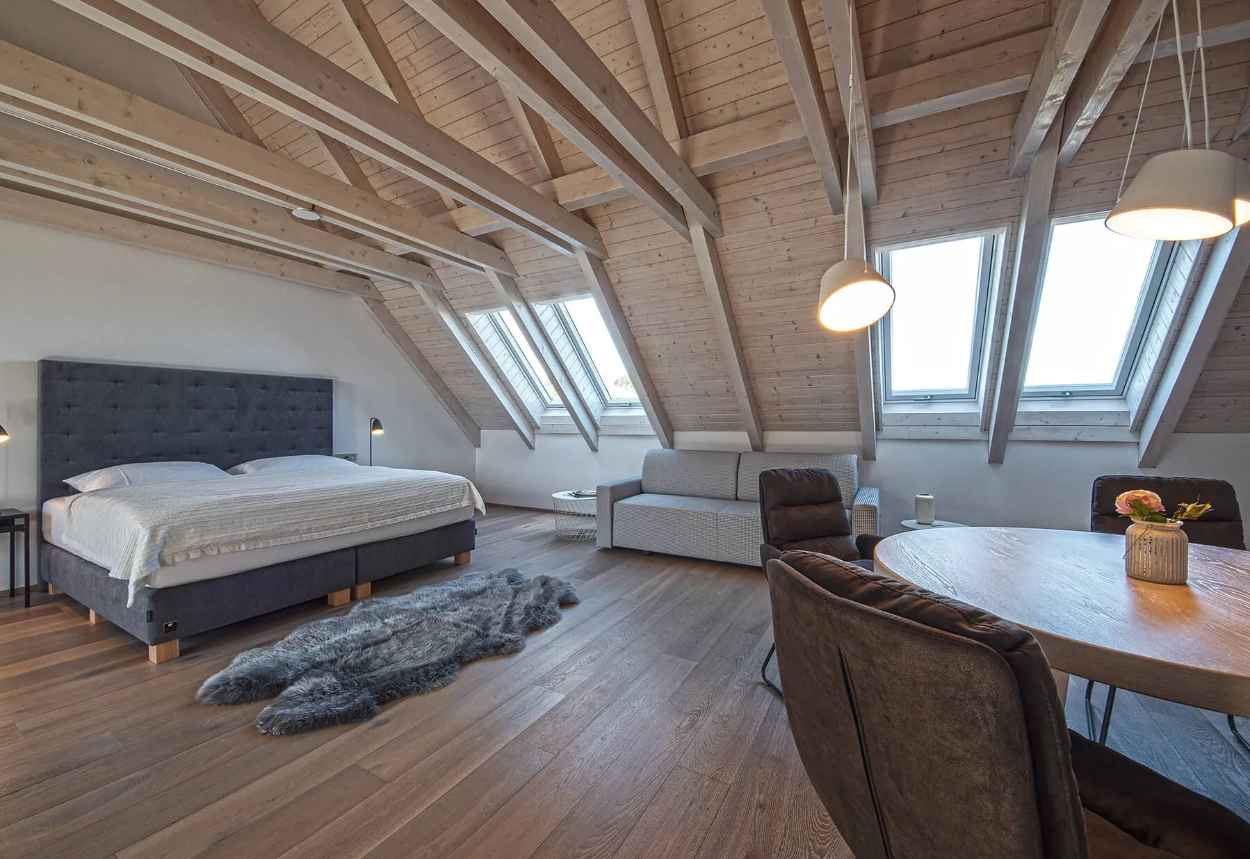 Moderní podkrovní ložnice s jídelním koutem, kde dominuje masivní dřevěná podlaha v kouřovém odstínu a přiznané trámové stropy s integrovanými střešními okny