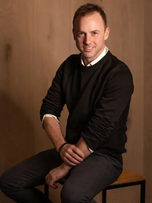 Portrét zakladatele společnosti, Karla Hrdličky, sedícího v ateliéru na dřevěné stoličce před stěnou s přírodním dřevěným obložením.