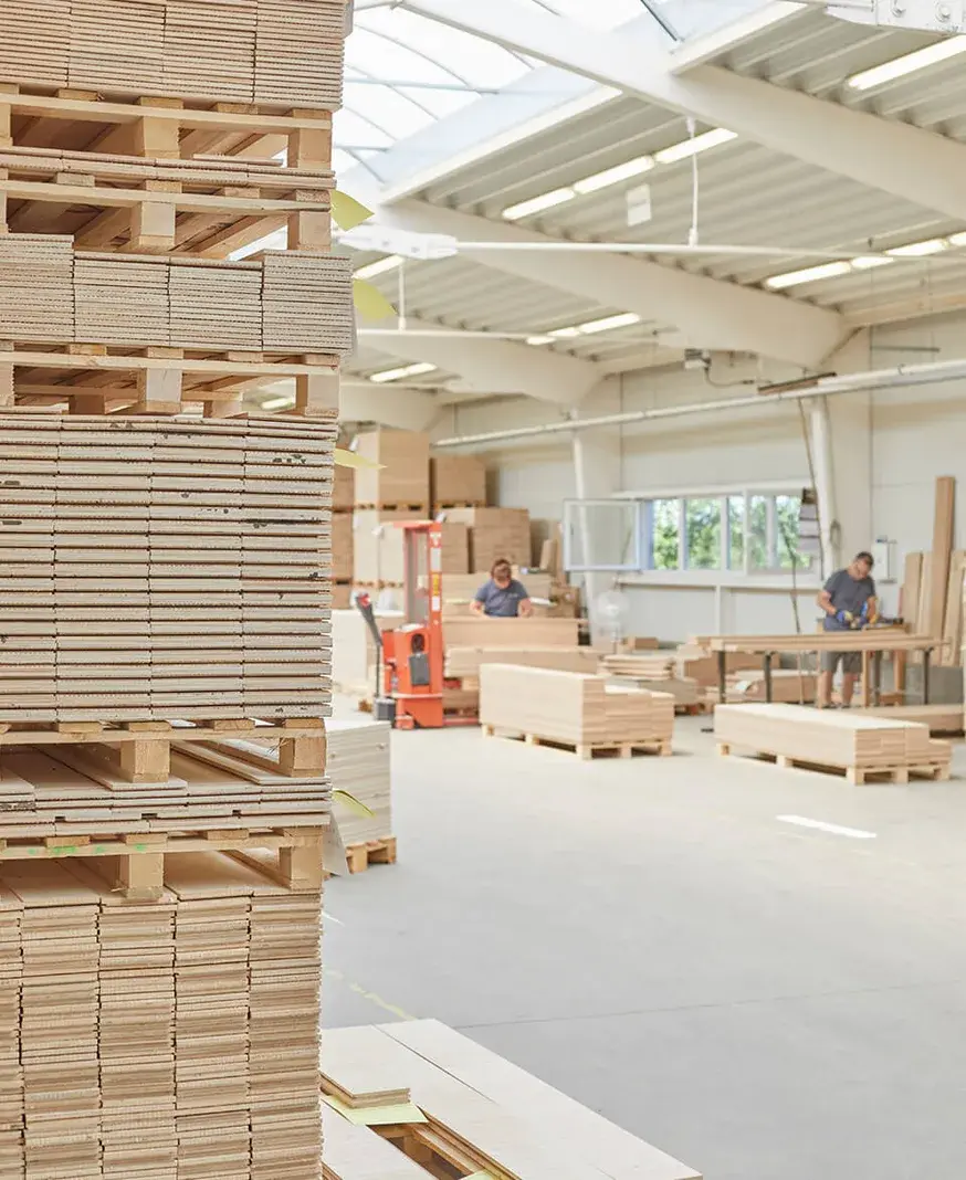 Pohled do prosvětlené výrobní haly s naskládanými paletami dřevěných lamel, kde pracovníci v pozadí zpracovávají dubové dřevo pro výrobu podlah.